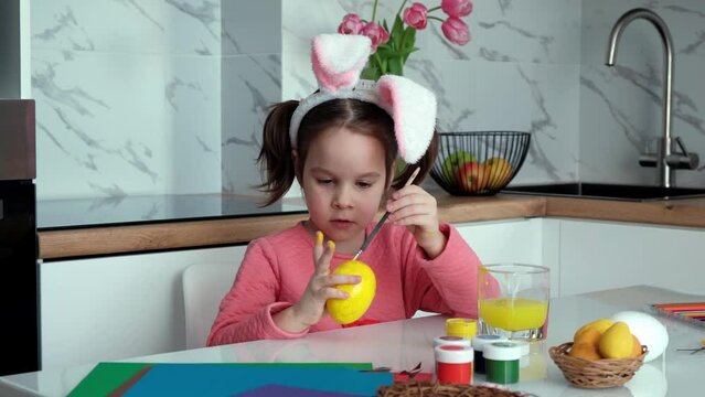 A dark-haired little girl with bunny ears and a pink dress paints Easter eggs at home in the kitchen.Happy Easter concept.