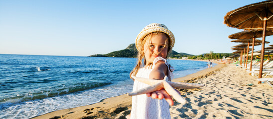 Cute little girl with straw hat show sea star on sand beach. Child at summer vacation.