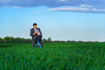 Naklejka na ściany i meble businessman dressed in a business suit, poses with bicycle in green grass field and reads documents, beautiful nature in spring, business concept