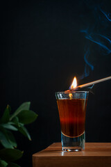 Ignition of cocktail made of hard liquor mixed with grenadine syrup served in shot glass on brown wooden bar counter against black wall with green plant and smoke. Vertical image with copy space
