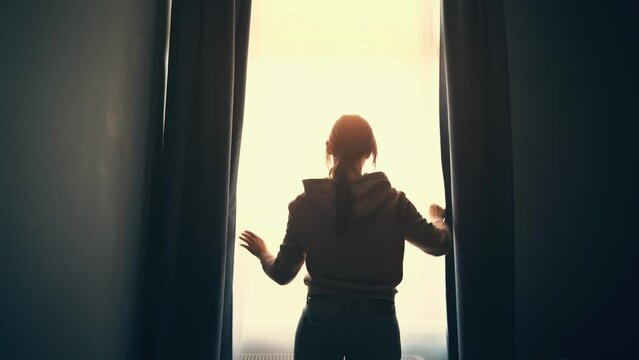 Girl opens window curtains in dark room to sunlight, rear view. New day and hope concept.