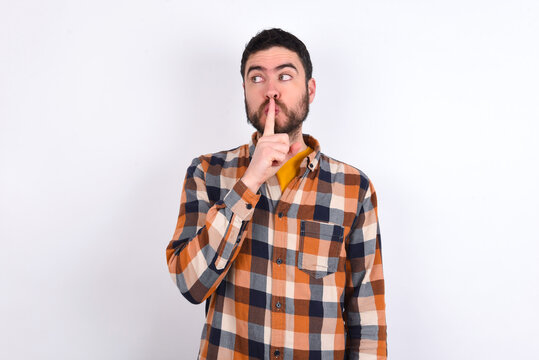 young caucasian man wearing plaid shirt over white background makes silence gesture, keeps index finger to lips makes hush sign. Asks not to share secret.