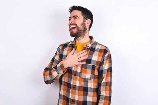 Joyful young caucasian man wearing plaid shirt over white background expresses positive emotions recalls something funny keeps hand on chest and giggles happily.