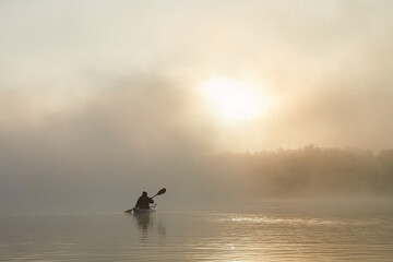 Fototapeta premium Man in kayak paddling through dense fog in wilderness area at autumn sunrise