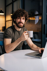 Vertical portrait of positive curly young businessman holding in hand cup with morning coffee sitting at table with laptop computer, looking at camera, in kitchen with modern interior, remote office.