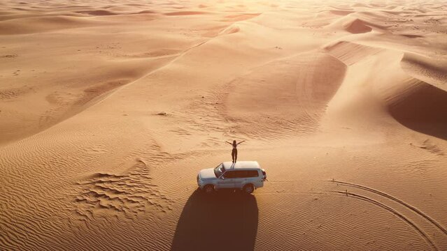 Aerial view of female traveller stands on the car's roof and rises her hands on the sunset in the desert.