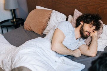 High-angle view of tired bearded young man sleeping peacefully lying on side in large comfortable double bed under white blanket with head on soft pillow, having pleasant good dream at home.