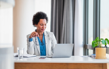 Smiling young adult mature African American businesswoman working on a laptop at her desk in a bright modern office holding her glasses.