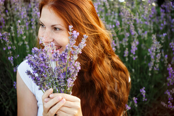 Naklejka premium Beautiful smiling woman in lavender field. Beauty portrait of girl with lavender bouquet flowers. Close up face with freckles and red long hair