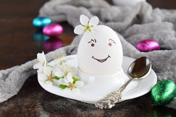 Smiling white egg and flowers on clay plate. Happy Easter card. Holidays breakfast concept. Festive table place setting decoration with blossom, bunny, chocolate eggs.