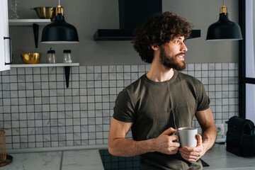 Portrait of handsome pensive young man holding in hand cup with morning coffee, thoughtful looking out window, standing in kitchen with modern light interior, enjoying fresh warm drink at home office.