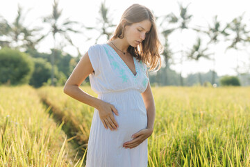 Pregnant woman stand at the field with green grass and embrace her belly. Sunshine summer vibes
