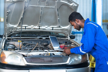 Car mechanic using laptop while checking car engine or circuit at garage - concept of professional, technology, expertise and maintance service.