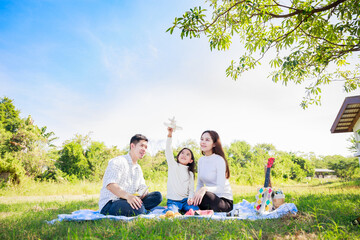 Happy family picnic. Asian parents (Father, Mother) and daughter playing the toy airplane and have enjoyed ourselves together while picnicking on picnic cloth in green garden in the sunshine day
 - obrazy, fototapety, plakaty
