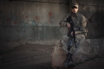 Fototapeta premium a young Ukrainian soldier in camouflage uniform, wearing tactical goggles and a cap holding a black machine gun inside a destroyed building. defender of the motherland with weapons