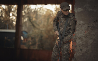 Naklejka premium a young Ukrainian soldier with combat experience in a gray camouflage uniform, wearing tactical glasses and a cap with a black machine gun in his hands peeks around the corner in search of the enemy