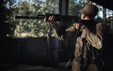 Fototapeta premium a young soldier defender of his country in a camouflage gray uniform, wearing tactical black glasses and a cap with a machine gun in his hands inside a large authorized building