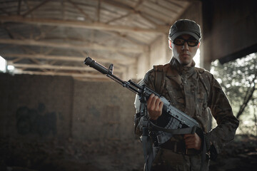 fearless soldier defender of the motherland in a camouflage protective uniform, in tactical black glasses and a cap with a machine gun in his hands stands inside a large authorized building