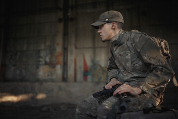 Obraz premium a soldier with a machine gun in a gray camouflage uniform wearing a cap and tactical black glasses sits on a concrete floor inside a large destroyed building