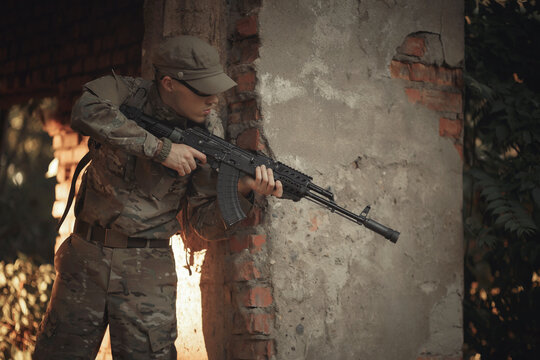 a soldier with combat experience in a gray camouflage uniform, wearing tactical glasses and a cap with a machine gun in his hands, peeks around the corner in search of the enemy