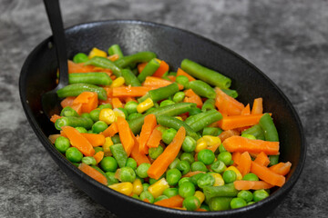 Mixed vegetables, peas, carrots, green beans and corn, in a black dish.. On a black stone background