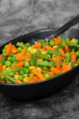 Mixed vegetables, peas, carrots, green beans and corn, in a black dish.. On a black stone background