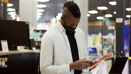 African American man in mask is choosing a new mobile phone in a shop, checking how it works