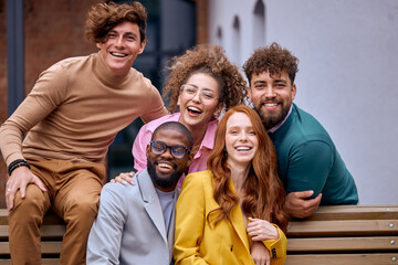Portrait of successful group of people enjoying free time together outdoors. Guys and ladies in stylish formal outfits after working in ecommerce business office, laughing,sitting on bench
