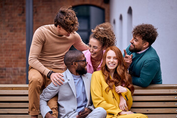 Five co-workers have rest outdoors in downtown, sitting on bench laughing, relaxed men and women get pleasure by spending time together, enjoying meeting, multiracial mixed race group