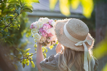 Beautiful blonde in a green summer garden. A girl in a straw hat with a bouquet of white peonies and pink carnations.