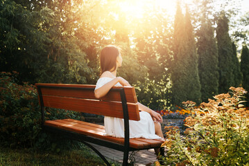 Young brunette woman in white summer dress resting, relaxing in flower garden outdoors. Back view of an elegant lady sitting on bench and enjoying sunset - obrazy, fototapety, plakaty