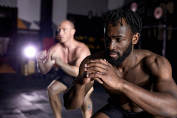 Obraz na płótnie Canvas two an athletic men doing box jump exercise. Cross fit, sport and healthy lifestyle concept. African and caucasian shirtless sportsmen engaged in sport, concentrated and motivated. focus on black man