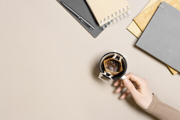 Office desk table, woman work and hand hold cup with drip coffee bag, planner, copybooks, notebooks on beige background. Paper bag drip coffee filter for brewing ground coffee, top view