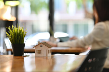 Cropped view of a house model and a key on an office table with young Asian woman working in the office in the background, for real estate business and finance concept.