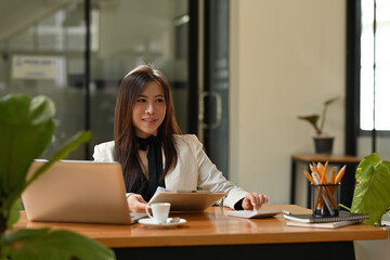 A portrait of a young pretty Asian woman sitting in the office using a laptop, calculator, working on data, charts and documents on the table, for business, finance and technology concept.
