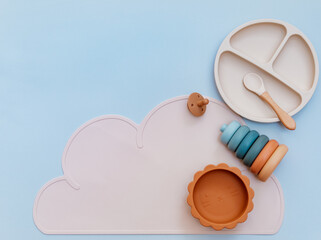 Pastel silicone collection of tableware, bibs, accessories, Montessori toys for children on blue background. Baby accessories, tableware concept.