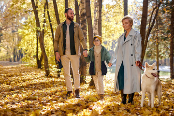 Fototapeta premium caucasian family with child and dog on walk in autumn forest, lead dog on a leash. Family walk with white dog in park. attractive man, woman and kid boy spend time together, love their pet