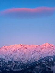 Morgenroth schittert roze in de noordelijke Alpen in de winter, verlicht door de zonsopgangzon op het bergoppervlak. De wolken die in de lucht zweven zijn ook roze.