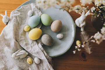 Fototapeta na wymiar Happy Easter! Easter eggs on plate with bunny figurines, linen napkin, cherry blossoms on rustic table. Natural dyed colorful eggs and spring flowers. Countryside still life, top view