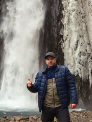 A man shows a cool gesture while standing at an icy stormy waterfall flowing down wet rocks