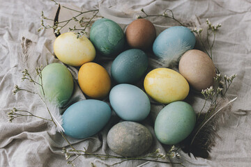 Happy Easter! Easter eggs on rustic linen napkin with tender flowers flat lay. Stylish natural dyed pastel colorful egg, spring flowers and feathers composition. Rustic still life
