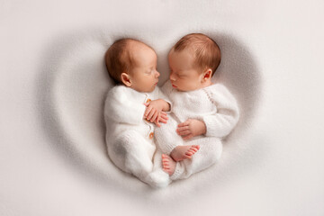 Tiny newborn twin boys in white bodysuits on a white background. Newborn twins sleep next to their brother on the background of the heart. Two newborn twin boys hugging each other.
