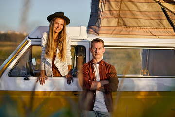 Portrait of young caucasian couple standing by yellow mini van looking at camera, posing, woman peep out from window, man with arms folded, travellers on trip in nature at summer evening