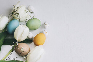 Happy Easter! Stylish Easter eggs and tulips border on rustic white wooden background with space for text. Natural colorful dyed eggs and spring flowers composition. Greeting card