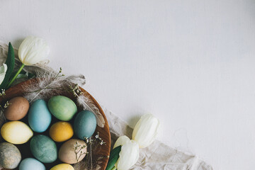 Stylish Easter eggs in wooden plate  and tulips on rustic white background. Happy Easter! Natural dyed colorful eggs and spring flowers composition on rustic table. Flat lay with space for text