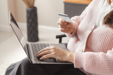 Fototapeta premium Woman makes a purchase on a laptop and pays with a credit plastic card. Millennial smiling woman 30 years old sits in a comfortable chair in a cozy home and does online shopping or orders delivery