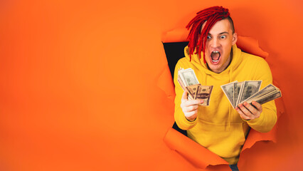 Young handsome man with money sticks out of hole of orange background. Stylish hipster with dollar bills.