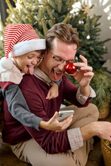 Joyful millennial father having fun laughing with adorable little kid son, taking photo on smartphone, sitting near decorated Christmas tree, enjoying festive winter holiday New Year celebration time