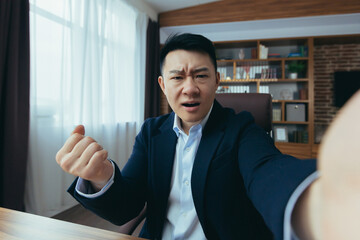 Fototapeta premium Angry Asian businessman boss talking on video call, using phone, working in classic office, arguing with employee, looking at phone camera
