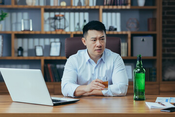 Asian businessman fails, man in despair drinks hard liquor, sitting late at the table in the office, depressed and hopeless